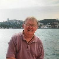 Photo of Sam Grant in front of a European or Middle Eastern lake scene on a cloudy and sunny day. He is wearing glasses and a light burgandy polo