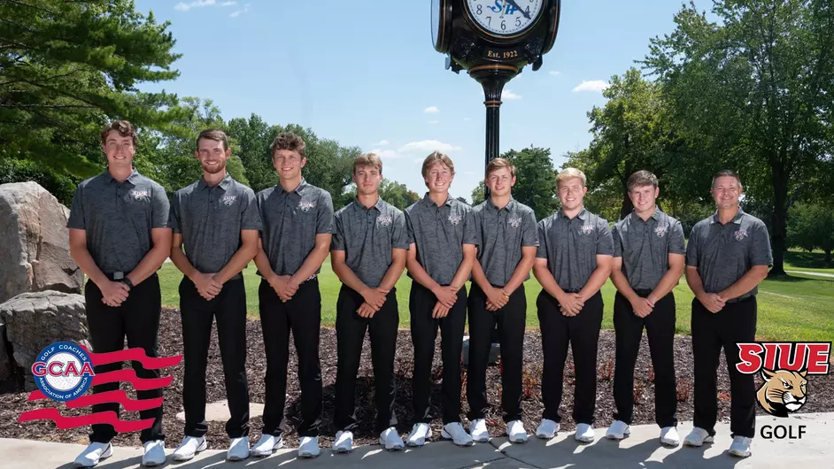 Group photo of the SIUE men's golf team. They are posing outside of an antique clock tower on a sunny day. It appears to be a golf course behind them.