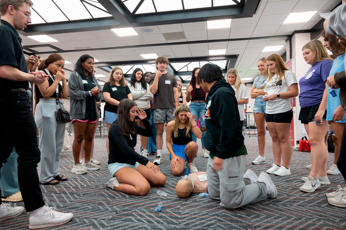 group of students standing in a circle observing two girls practicing CPR on a mannequin