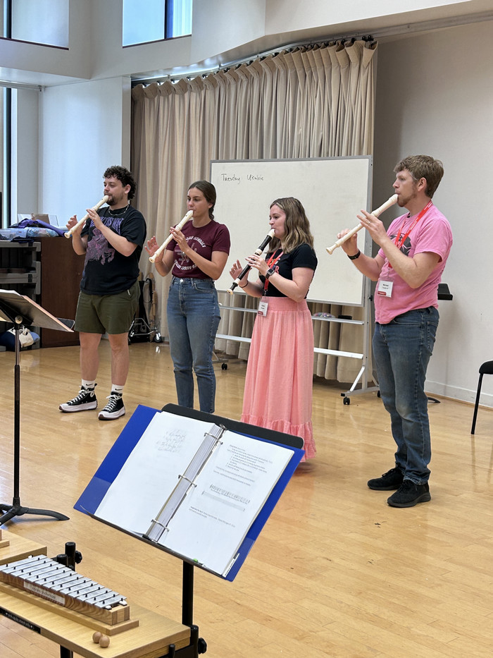 four adult class participants standing in a classroom, each playing large recorder-like flute instruments. They appear to be sharing an amusing moment with their smiles and posture. There is a music stand facing the camera.