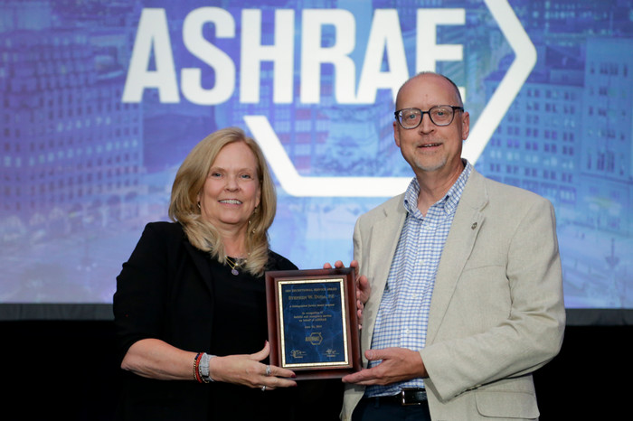 ASHRAE President Ginger Scoggins and SIUE's Stephen Duda posing for a photograph in front of an ASRAE presentation panel with "ASRAE" displayed behind them. They are holding up a rectangular, framed metal plaque about the size of a standard piece of paper. It is the Exceptional Service Award. 