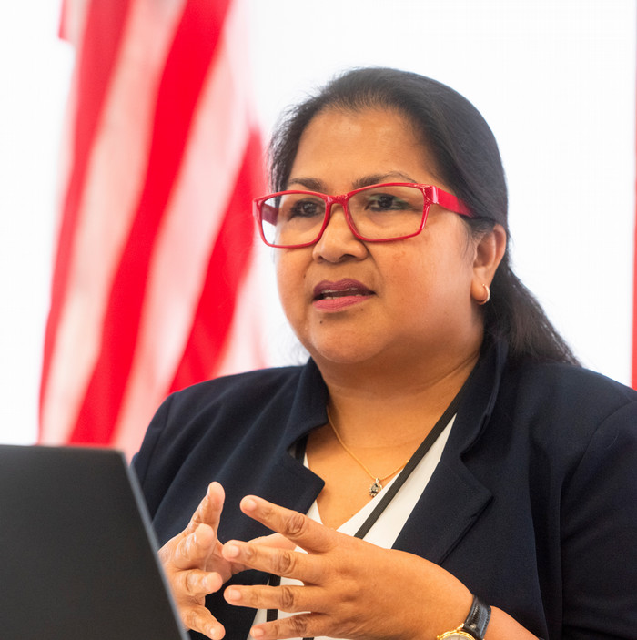 photo of Joey James speaking at a lecturn, the red and white stripes of the American flag out of focus behind her, her hands are clasped in front of her chest