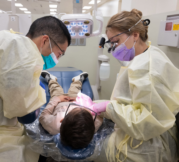 two dental students in pale yellow scrubs leaning over a chair looking into the mouth of their child patient 