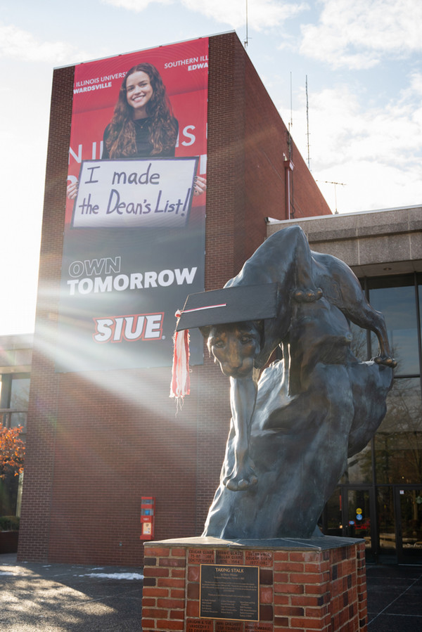 Cougar statue wearing a giant graduation cap and tassel on a cloudy day. In the background, there is a banner of a graduate holding a sign reading "I made the dean's list" hanging from the MUC